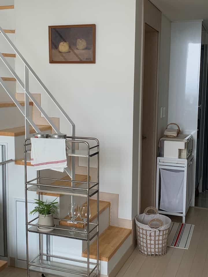 ホワイトと木目調が調和したロフトのキッチンスペース、鉄製トロリーとランドリーバスケットが配置され、清潔で実用的な雰囲気のインテリア