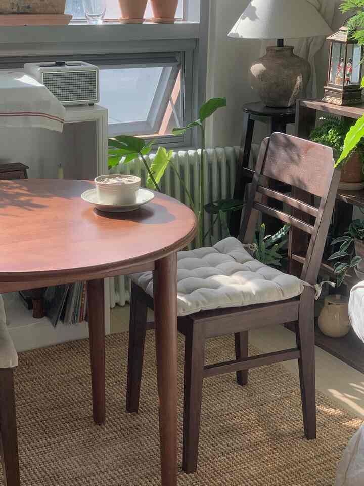 ウッドトーンのナチュラルな印象的な空間、丸テーブルとクッション付き木製椅子、窓辺の植物が配置されたカフェ風インテリア