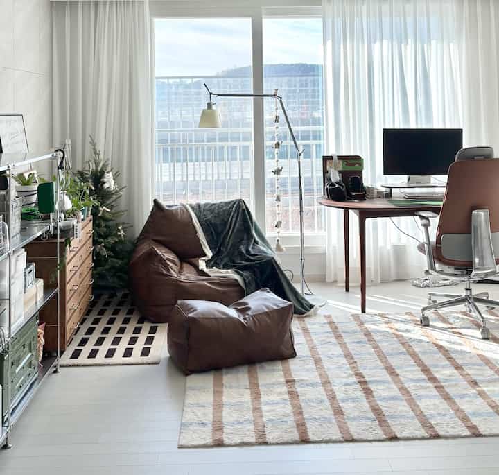 ホワイトトーンのリビングワークスペース、木製デスクとブラウンレザーのビーズクッションソファが配置されたシンプルで居心地の良いインテリア