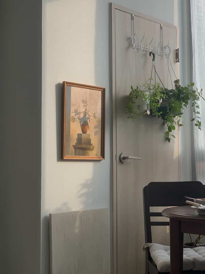 明るい木目調のドアにハンギングプランツを掛けたドアハンガーと、ヴィンテージの額縁が配置されたナチュラルで落ち着いたインテリア