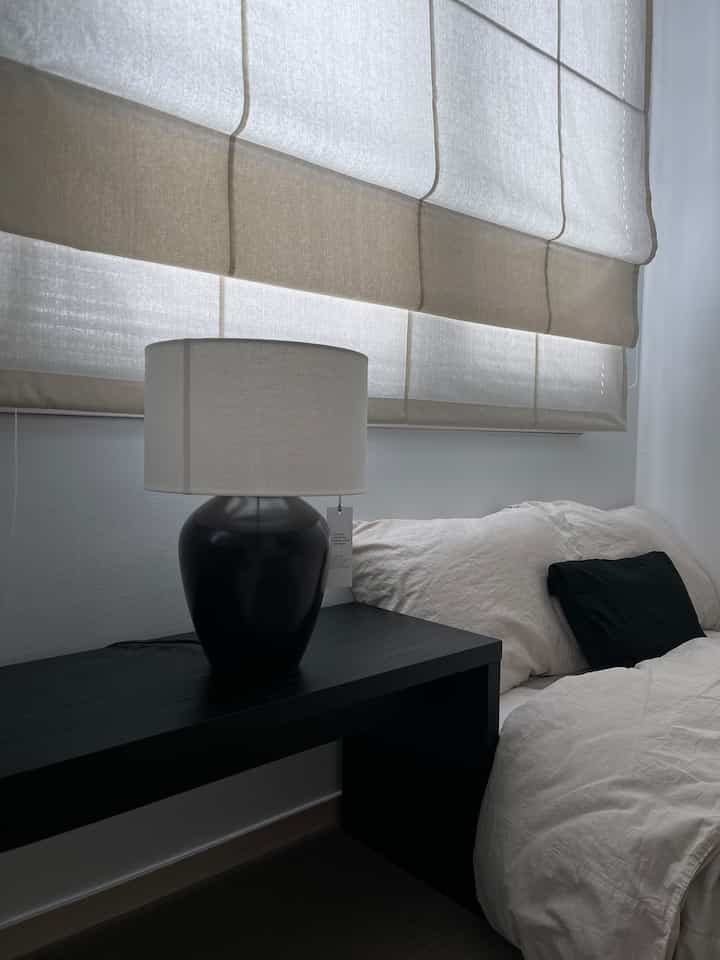 アイボリー色調の寝室、ローマンシェードと黒いナイトテーブルにテーブルランプが配置されたナチュラルでモダンなインテリア