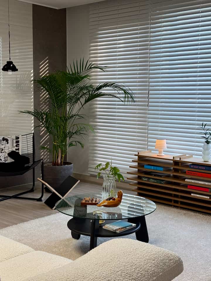 ナチュラルカラーと黒が調和したリビング、ガラスのコーヒーテーブル、アームチェア、植物が配置された雰囲気のある空間