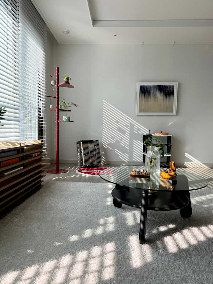 白い壁とカーペットのモダンなリビング、中央に透明ガラス天板の黒いコーヒーテーブル、窓際に赤いコートラックが配置された雰囲気のあるインテリア