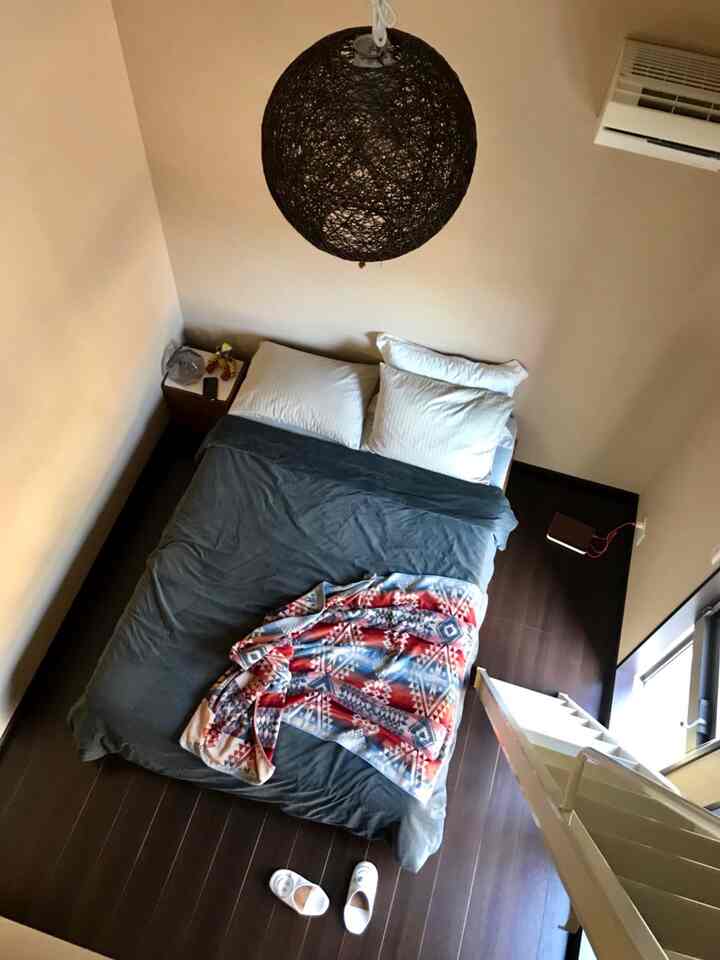 ベージュ色の壁とダークブラウンの床が印象的なロフト寝室、中央にカラフルな毛布をかけたベッドが配置された雰囲気のある空間