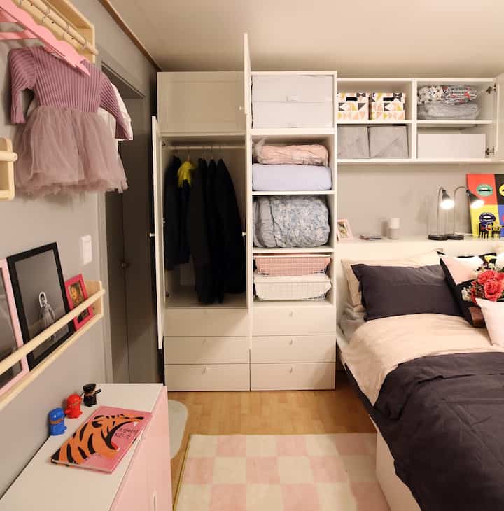 白とピンクの色調の寝室、整理された壁収納とクローゼット、子供の服やおもちゃが配置された雰囲気のある空間