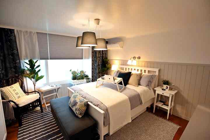 White and gray toned bedroom featuring central bed and bench, armchair on left, curtains and plants by the window creating a cozy atmosphere