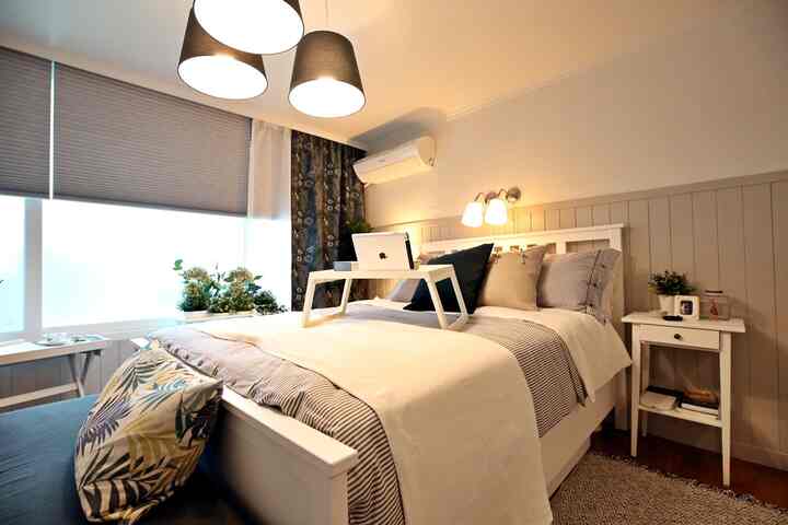 A cozy natural tone bedroom in white and beige, featuring a central bed, side table, and plants by the window