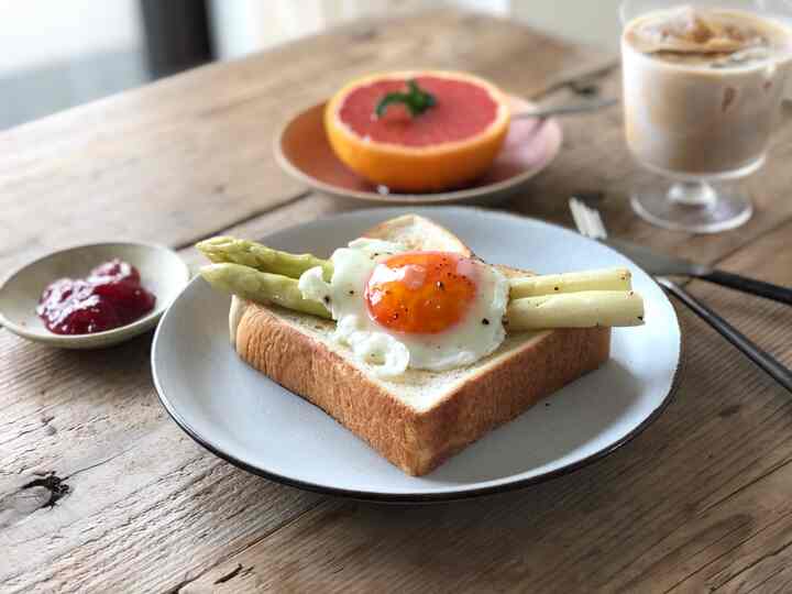 ナチュラルなブラウン調のキッチンダイニングテーブルにトーストと目玉焼き、アスパラガス、グレープフルーツ、アイスコーヒーが置かれた朝食の風景