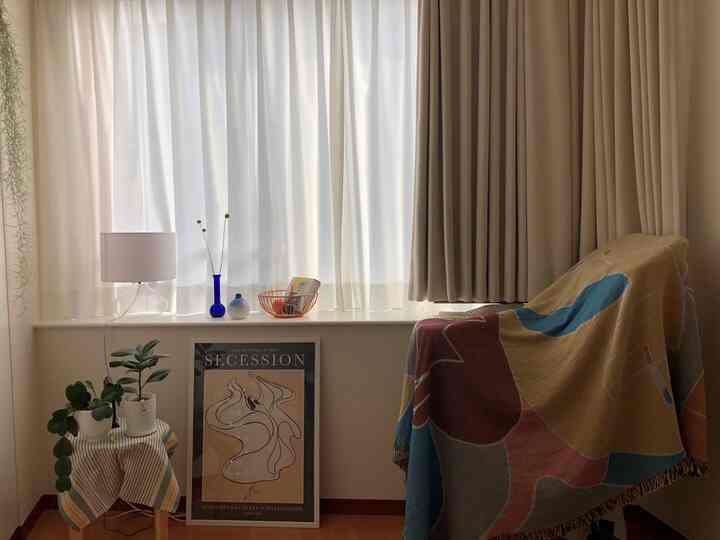 ベージュと白のナチュラルカラーのホームオフィス、サイドテーブルに植物とポスター、カラフルなブランケットがかかった椅子が配置された雰囲気のある空間