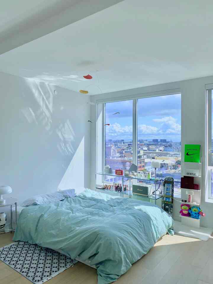 明るいホワイトの壁とライトブラウンの床がある寝室、薄いブルーの掛け布団カバー付きベッドと透明なガラス棚が配置された空間