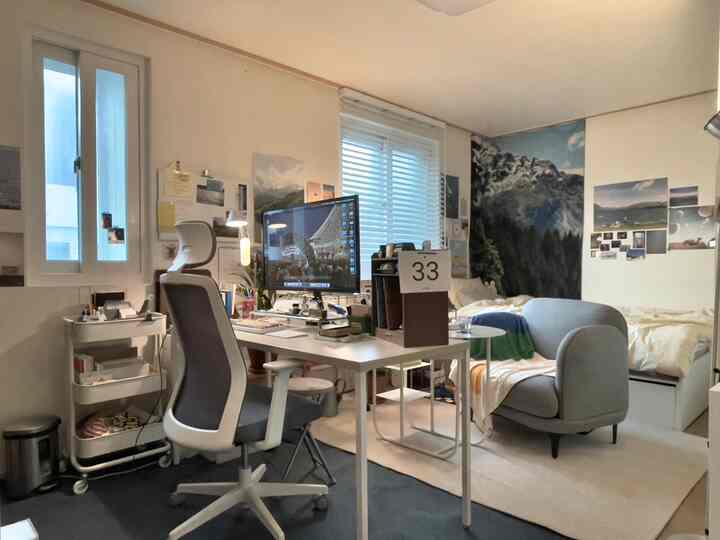 ホワイトとグレーのトーンが特徴の狭いワンルーム、ホームオフィスの机と椅子、モダンな寝室が調和した雰囲気のあるインテリア