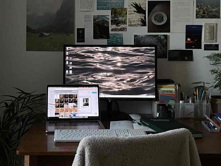 暖かみのある木目調とグレーのトーンが調和したホームオフィス、デスク上にはモニターとノートパソコン、壁にはポスターが配された落ち着いたワークスペース