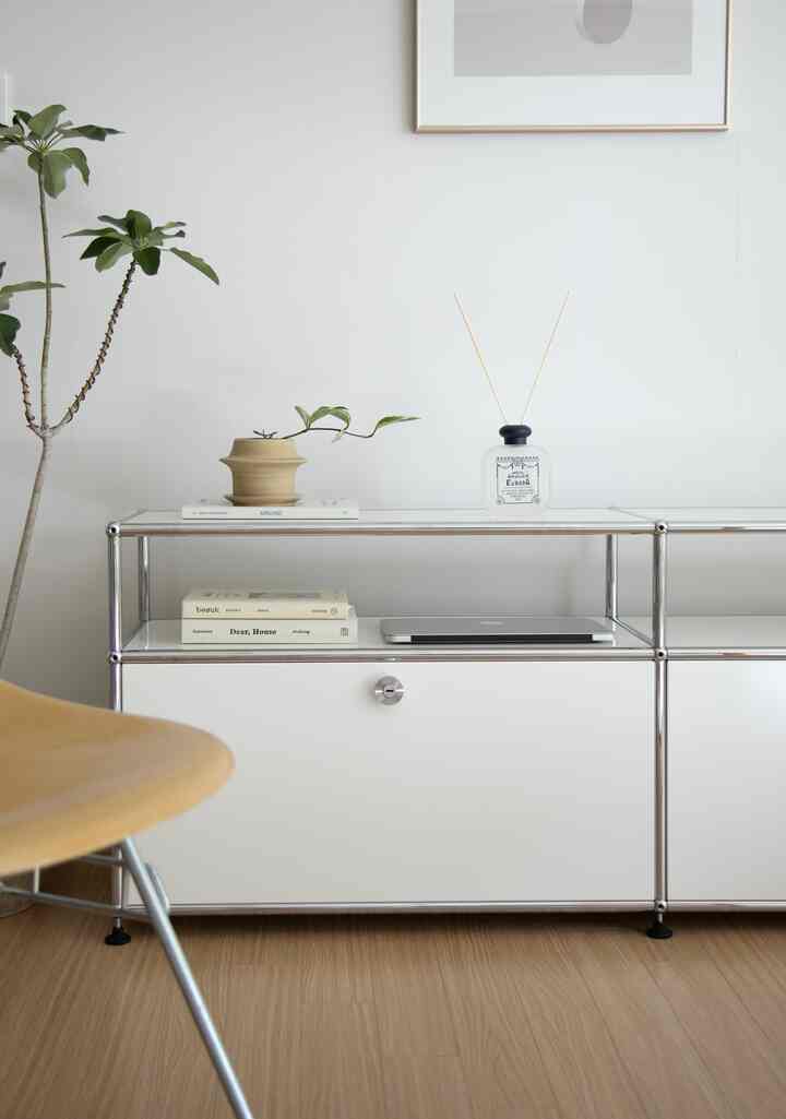 明るい白い壁と木目調の床のシンプルでミニマルなリビング、クローム棚、本とディフューザーが配置された雰囲気のあるインテリア