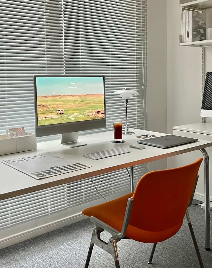 白とグレーのモノトーンでまとめられたコンパクトなホームオフィス、オレンジ色のオフィスチェアとアルミブラインドが配置された印象的な空間