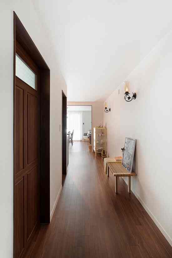 白い壁と木目調の床がある縦長の廊下、壁灯と木製ベンチが配置されたシンプルで自然な雰囲気の空間
