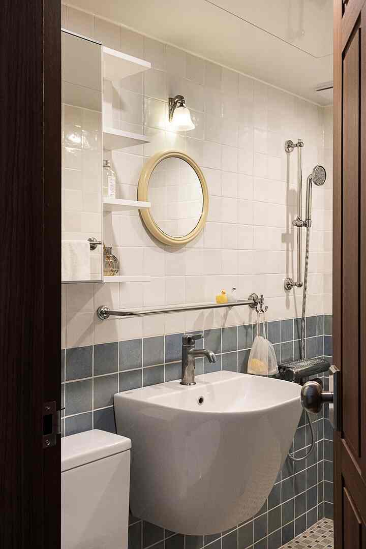 白と青のタイル張りのバスルーム、丸い鏡と洗面器、シャワーヘッド、飾り物が置かれた棚が配置された雰囲気のある空間