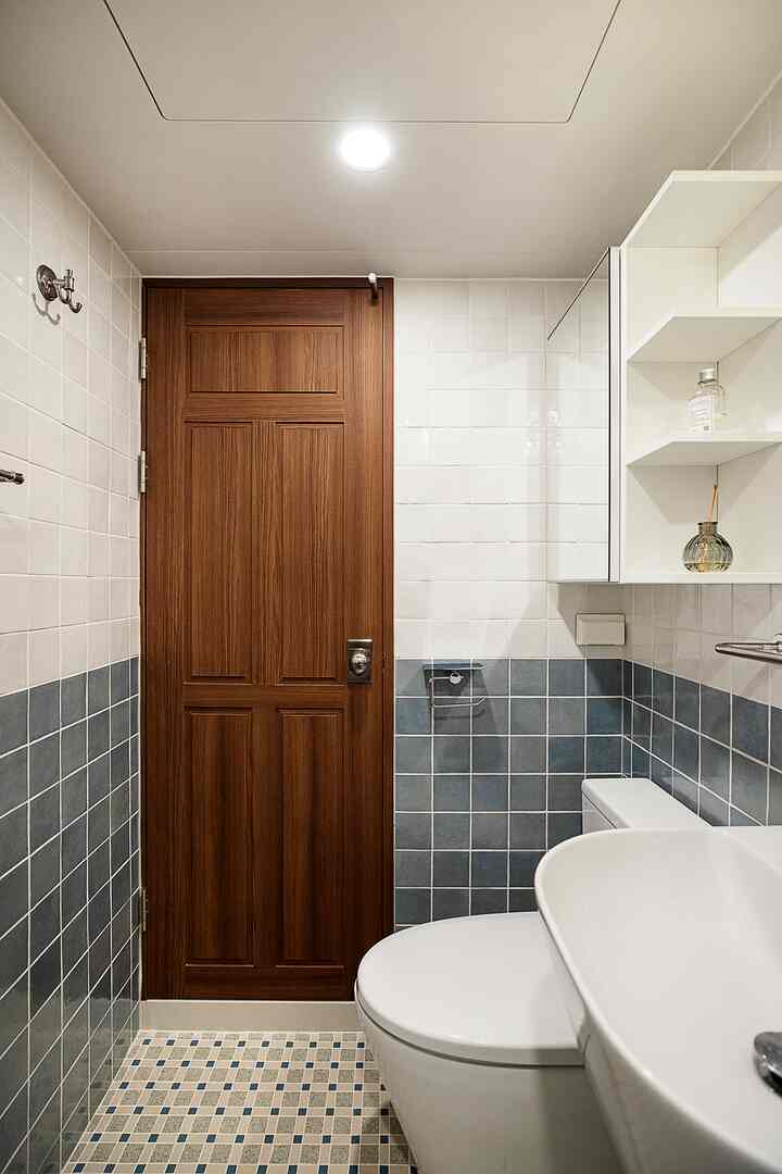 白と青のタイルが特徴の狭いトイレ、木製ドアと白い洗面台、トイレが配置された清潔な空間