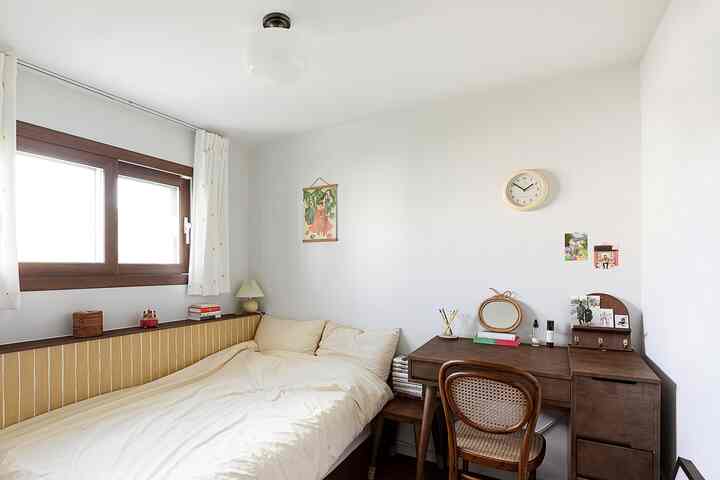 白い壁と天井が印象的な寝室、ブラウンの木製デスクとベッドが配置された自然で落ち着いた雰囲気のインテリア