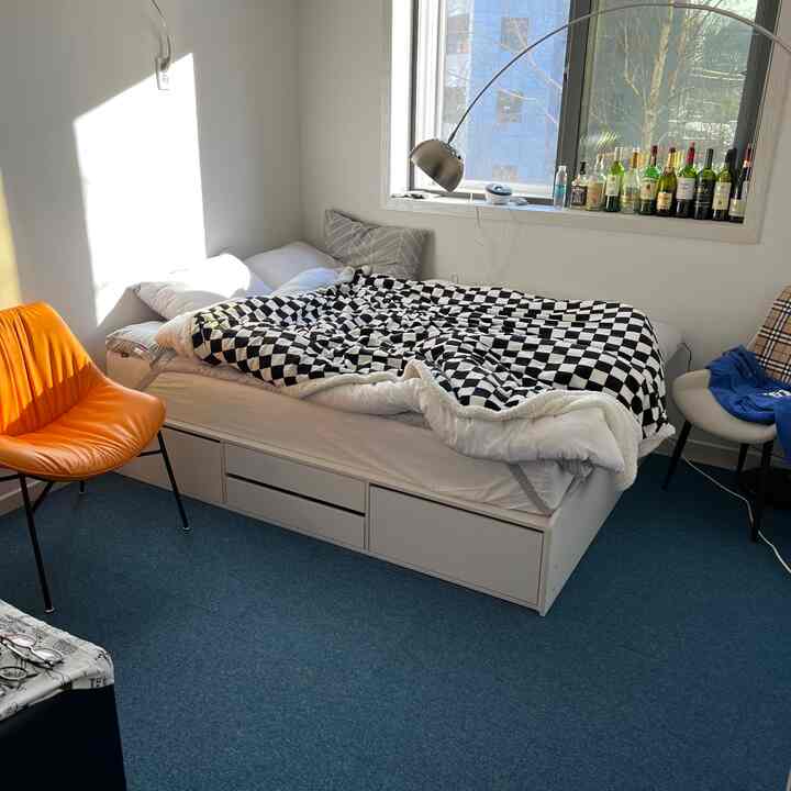 白い壁と青いタイルカーペットのある寝室、引き出し付きベッドとチェック柄の毛布、オレンジの椅子とシルバーカラーの照明が配置された雰囲気のあるインテリア。