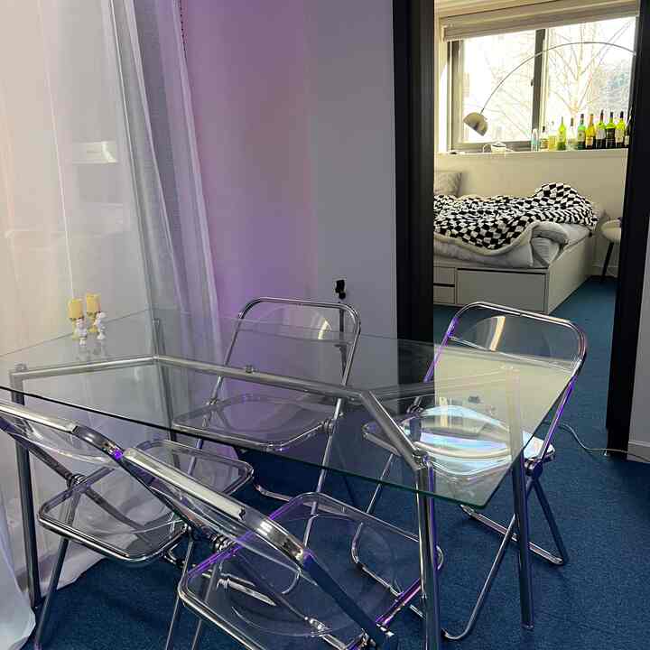 透明なガラスのダイニングテーブルとシルバーフレームの折りたたみ椅子が配置された、狭い空間のダイニングルームと寝室が見えるインテリア