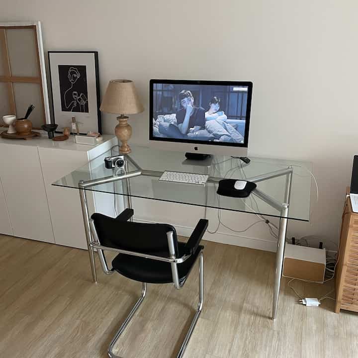 白い壁と木目調の床が調和したワンルームのホームオフィス、ガラスデスクと黒のアームチェアが配されたシンプルモダンな空間