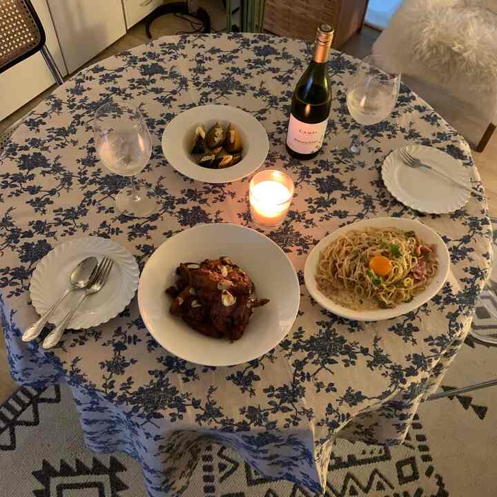 青い花柄のテーブルクロスがかかった丸いダイニングテーブルに皿、ワイングラス、料理、キャンドルが配置された雰囲気のあるホームパーティーの空間