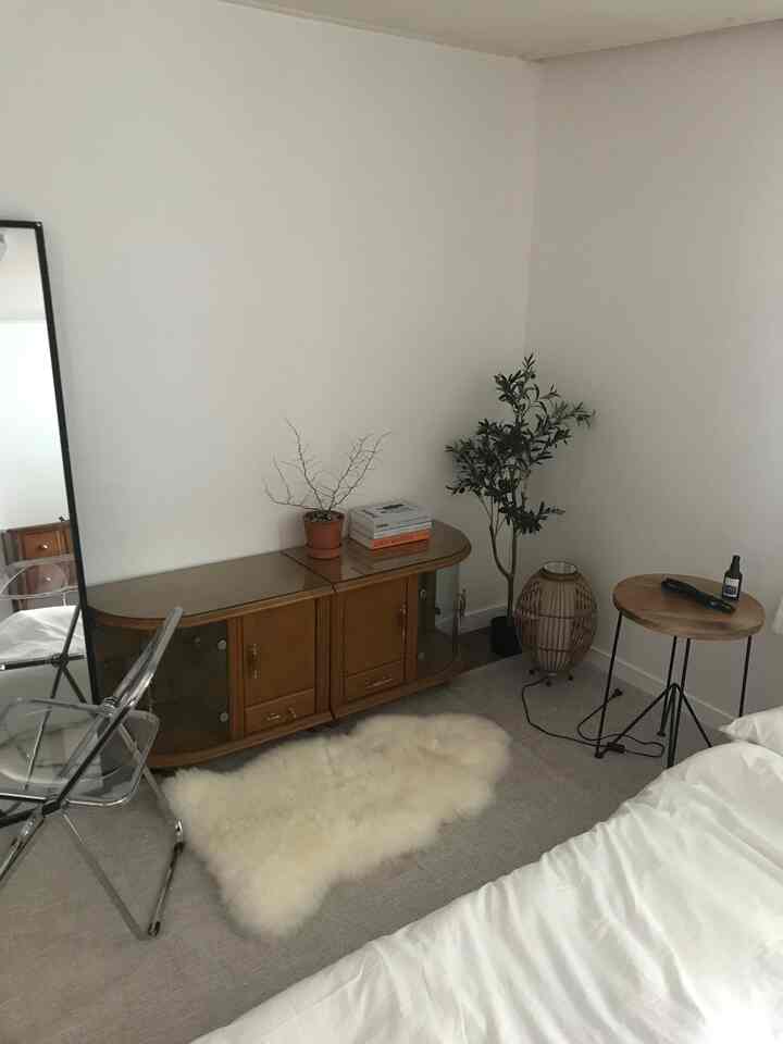 白い壁とナチュラルな木製家具がある狭い寝室、ベージュのラグと透明な椅子が配置されたミニマルな雰囲気のインテリア