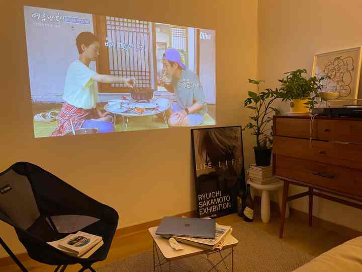 暖かいベージュと木目調が調和したコンパクトなリビング、プロジェクター画面とモダンな家具、植物が配置された雰囲気のあるインテリア