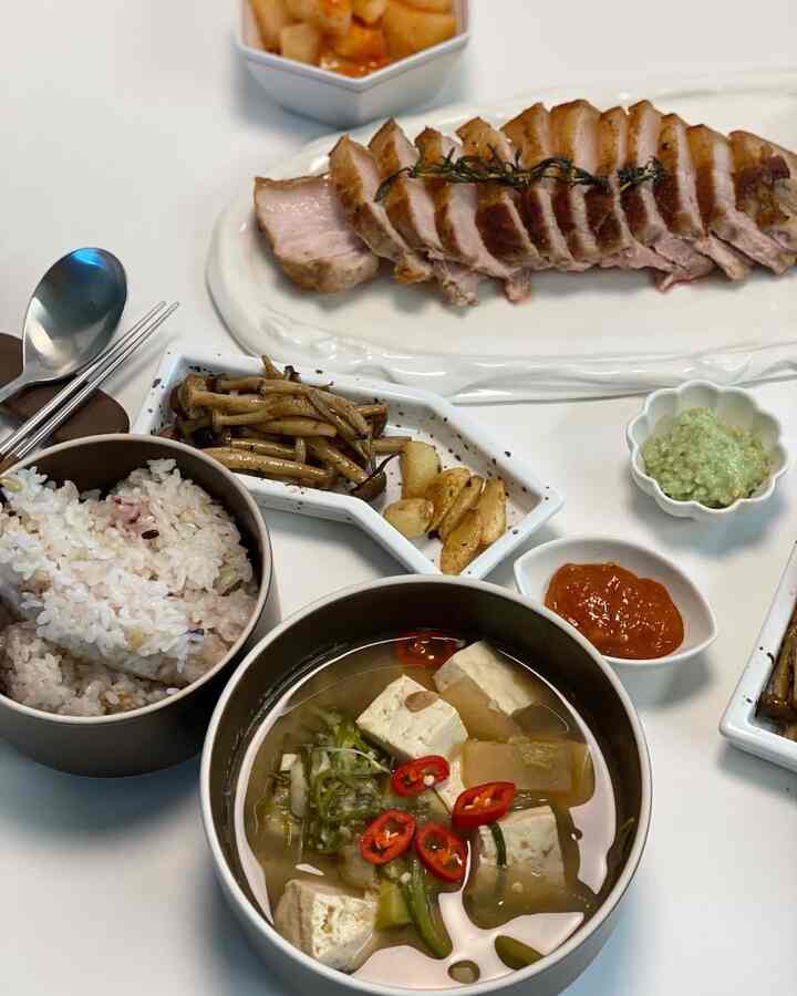 白いテーブルに味噌チゲの入ったボウル、ご飯の器、焼き肉の皿とおかずが並んだ食事風景