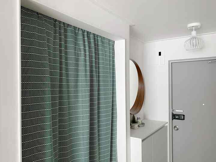 白い壁とグレーの玄関ドア、木目調の丸い鏡と柄のカーテンが配された実用的な玄関のインテリア