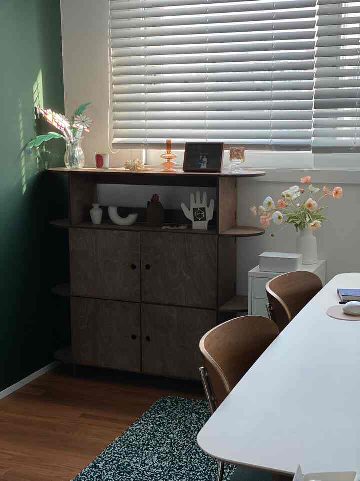 ダークグリーンの壁と木目調のキャビネットが配置されたナチュラルスタイルのリビング、白いダイニングテーブルと椅子が温かみのある雰囲気を演出。