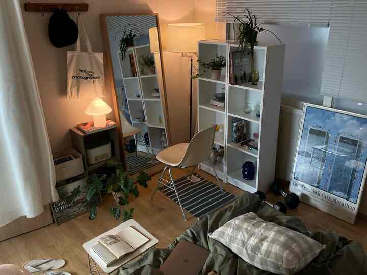 白い壁と木目調の床があるワンルームの寝室、ヴィンテージ照明と棚、植物が配置された雰囲気のあるインテリア