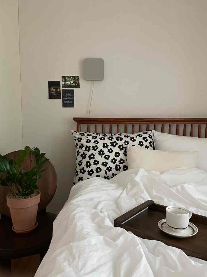 白とナチュラルな色調の寝室、木製のベッドフレームと白い寝具、左側に観葉植物と木製のサイドテーブルが配置された落ち着いた雰囲気のあるインテリア