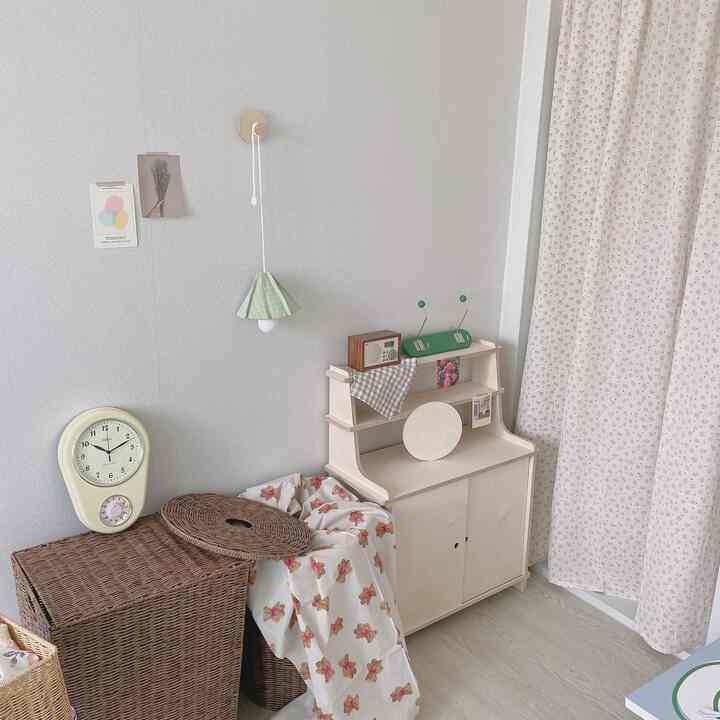 ホワイトの壁にラタンハンパーと木製キャビネット、花柄カーテンが配置されたナチュラルな狭い部屋