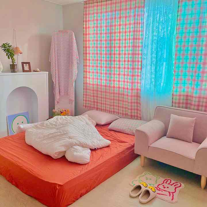 明るくナチュラルな色調の6畳の部屋、チェッカーカーテンとレースカーテン、低めのマットレスと小さなソファが配置された雰囲気のあるインテリア