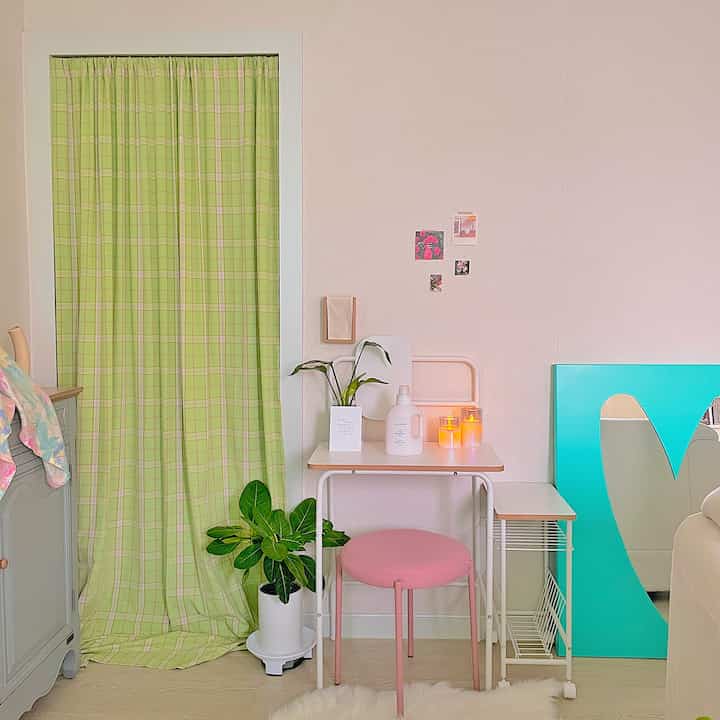 淡いグリーンのチェックカーテンとピンクのスツール、観葉植物が置かれたコンパクトで自然な雰囲気の空間
