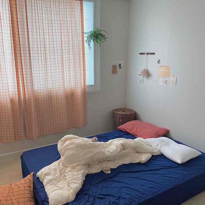 柔らかいベージュ色のカーテンと鮮やかなブルーのマットレスカバーが調和した居心地の良い寝室、白い寝具とラタンのハンパーがアクセントになっているインテリア