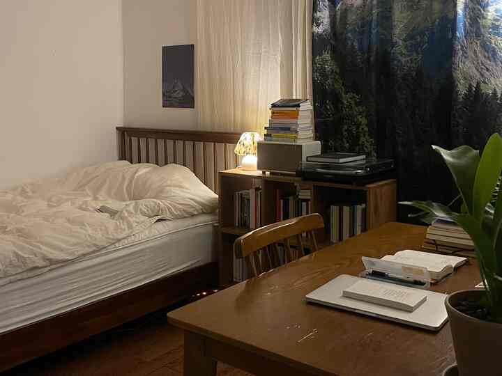 ブラウンの木製家具と白い寝具が調和したワンルームの寝室兼書斎、自然光とムードランプが温かみのある雰囲気を演出するインテリア