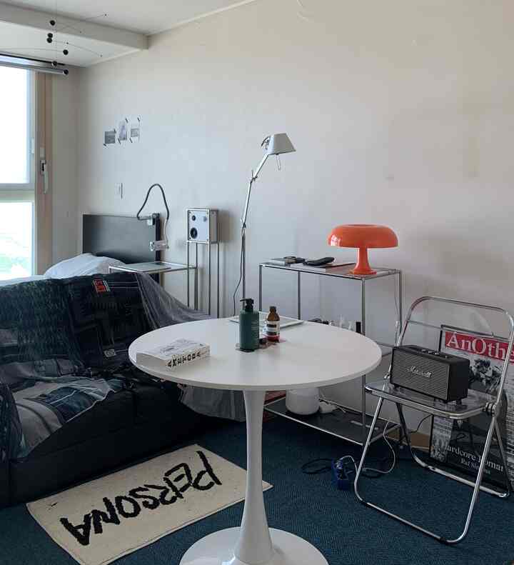 白と黒のトーンのワンルームリビングルーム、丸テーブルと黒いソファ、金属棚の上にオレンジのランプが置かれたシンプルな空間