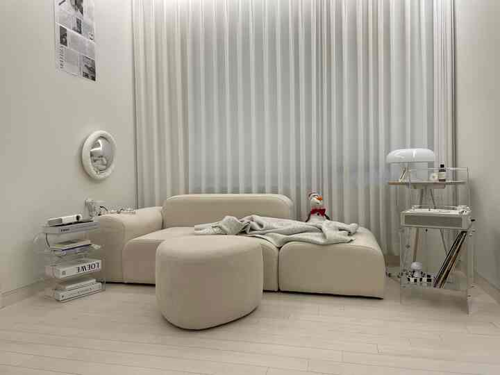 ベージュ調の二人暮らし向けリビングルームで、L字ソファとスツール、透明なコンソールテーブルが配置されたミニマルモダンなインテリア。