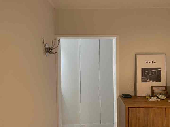 ベージュとウッドトーンのナチュラルカラーが映える寝室の玄関、壁にはコートハンガー、キャビネット上に額縁が配置されたシンプルで清潔感のあるインテリア