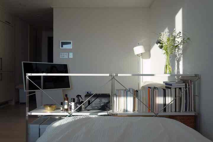 明るいホワイトトーンの寝室、ベッド背後の金属棚に本と花瓶が配置されたモダンな雰囲気のインテリア