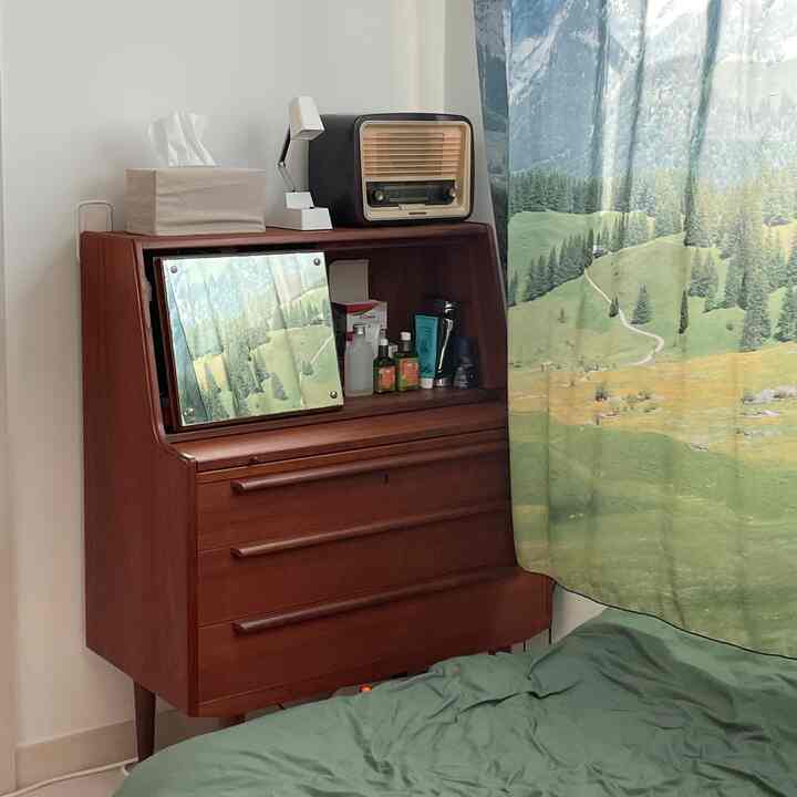 白い壁と木目調のヴィンテージ化粧台が配置された寝室、緑の寝具と自然を感じるカーテンが温かい雰囲気を演出