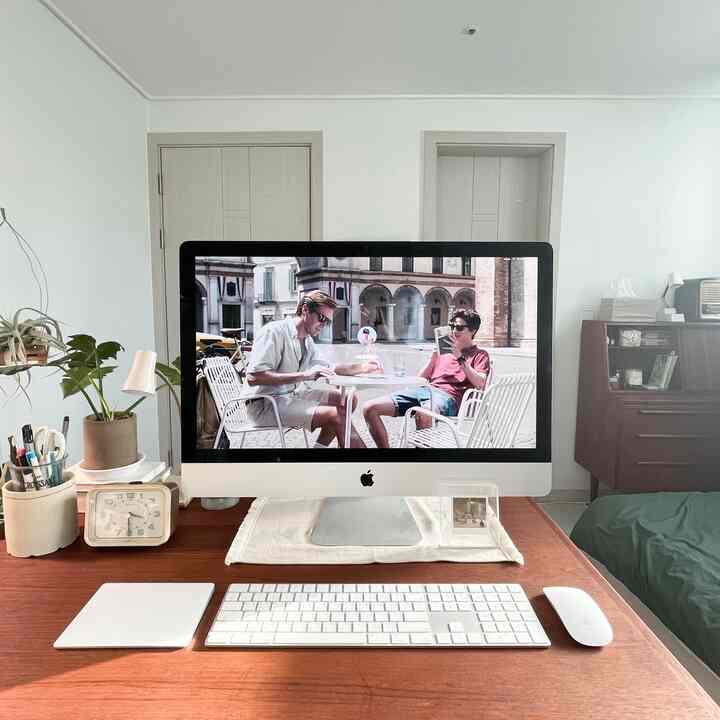 白い壁と木目調の机がある寝室兼ホームオフィス、Macコンピュータとベッドが配置された雰囲気のあるインテリア