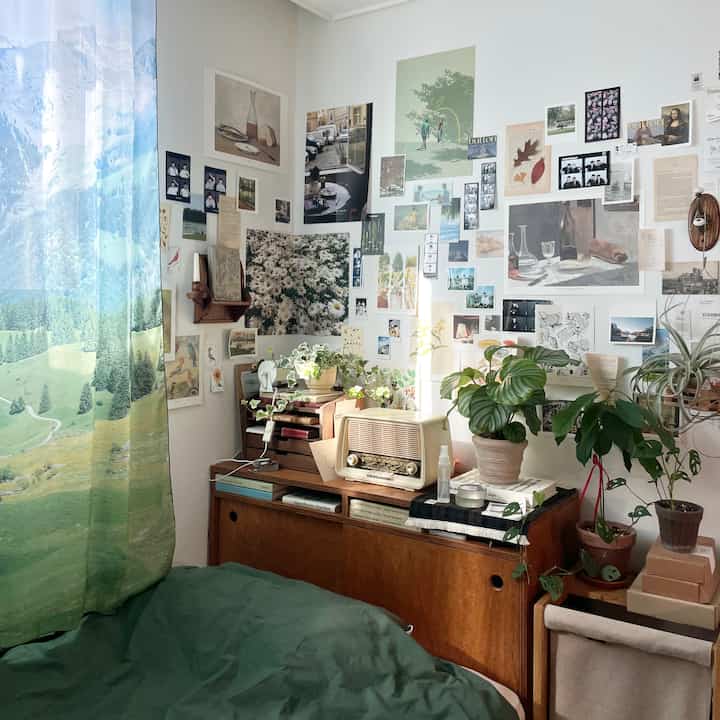 ナチュラルな木目調の家具と緑の植物が配置された6畳の部屋、壁一面に貼られた多様なアートポスターと印象的なカーテンのある空間