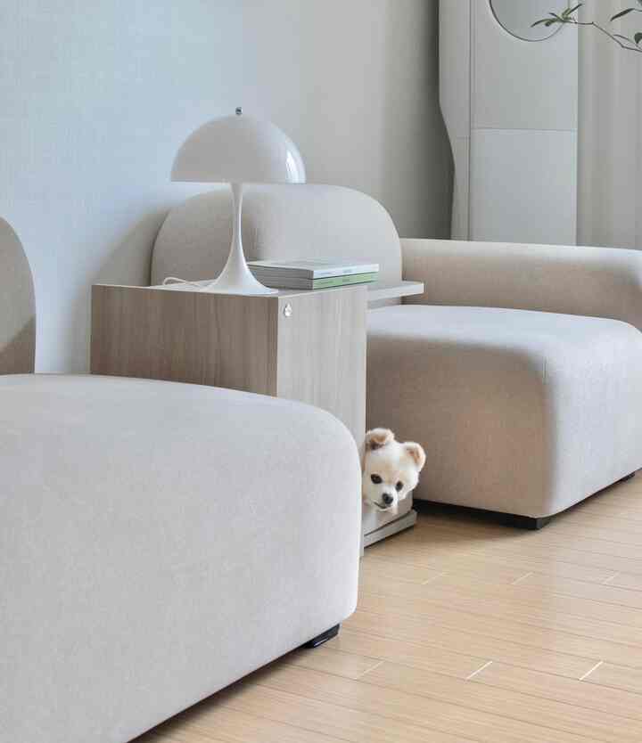 柔らかなナチュラルカラーのリビング、ベージュのソファと木目調のサイドテーブル、白いテーブルランプが置かれており、小さな犬がソファの間から顔をのぞかせている印象的な空間