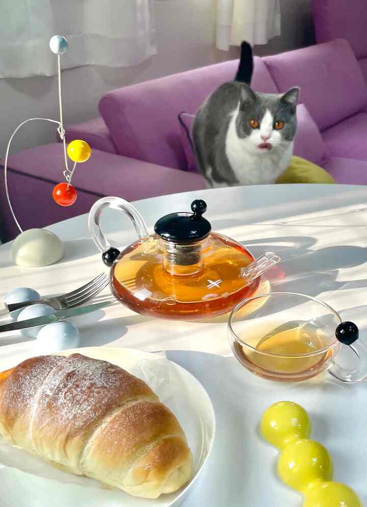 ピンク色のソファを背景にしたおうちカフェ空間、ソファに猫が座り、テーブルの上にはガラスのティーポットとクロワッサンが置かれ、温かみのある雰囲気を演出