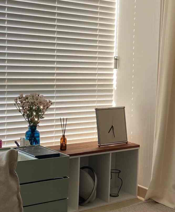 白い壁とブラインドのあるリビング、木目調の本棚と花瓶が置かれた雰囲気のあるインテリア