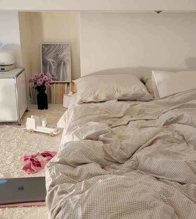 ベージュの色調で落ち着いた寝室、チェック柄の寝具とラグ、壁に額縁と照明が配置されたシンプルな空間