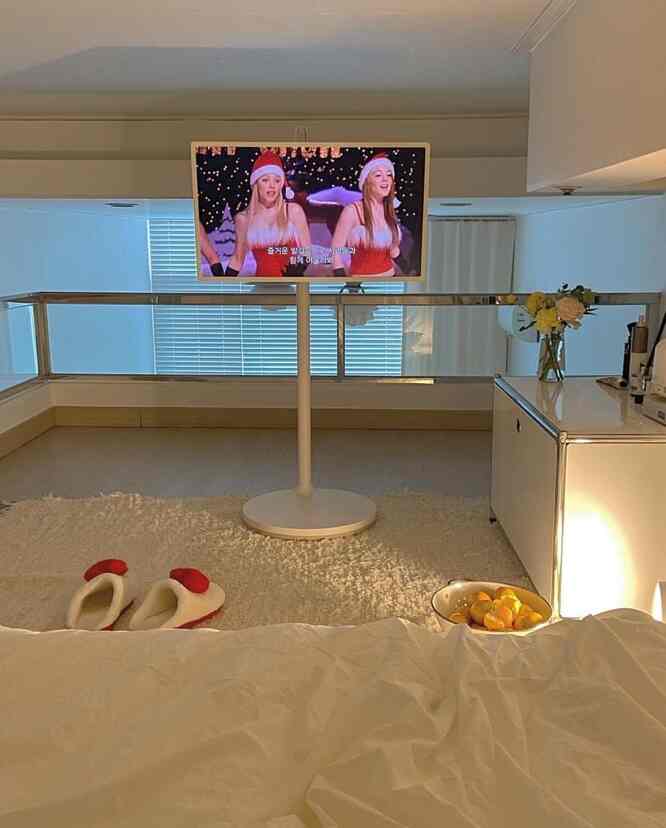 明るいホワイトトーンのリビングルーム、中央にテレビ台と壁掛けテレビが配置され、柔らかなラグと寝具が調和したシンプルなインテリア